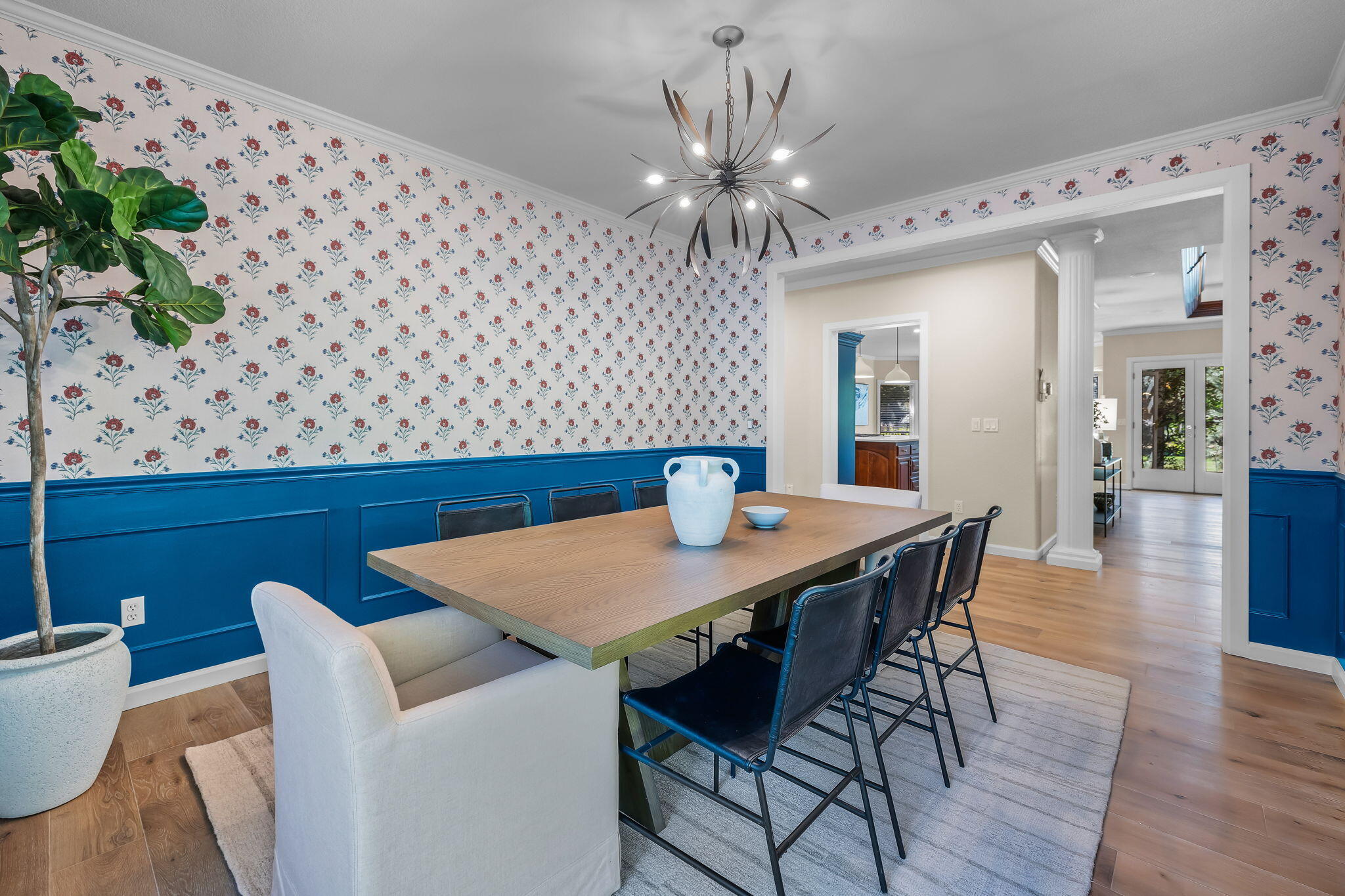 Dining room with floral wallpaper, blue wainscoting, and a wood table beneath a sculptural chandelier.