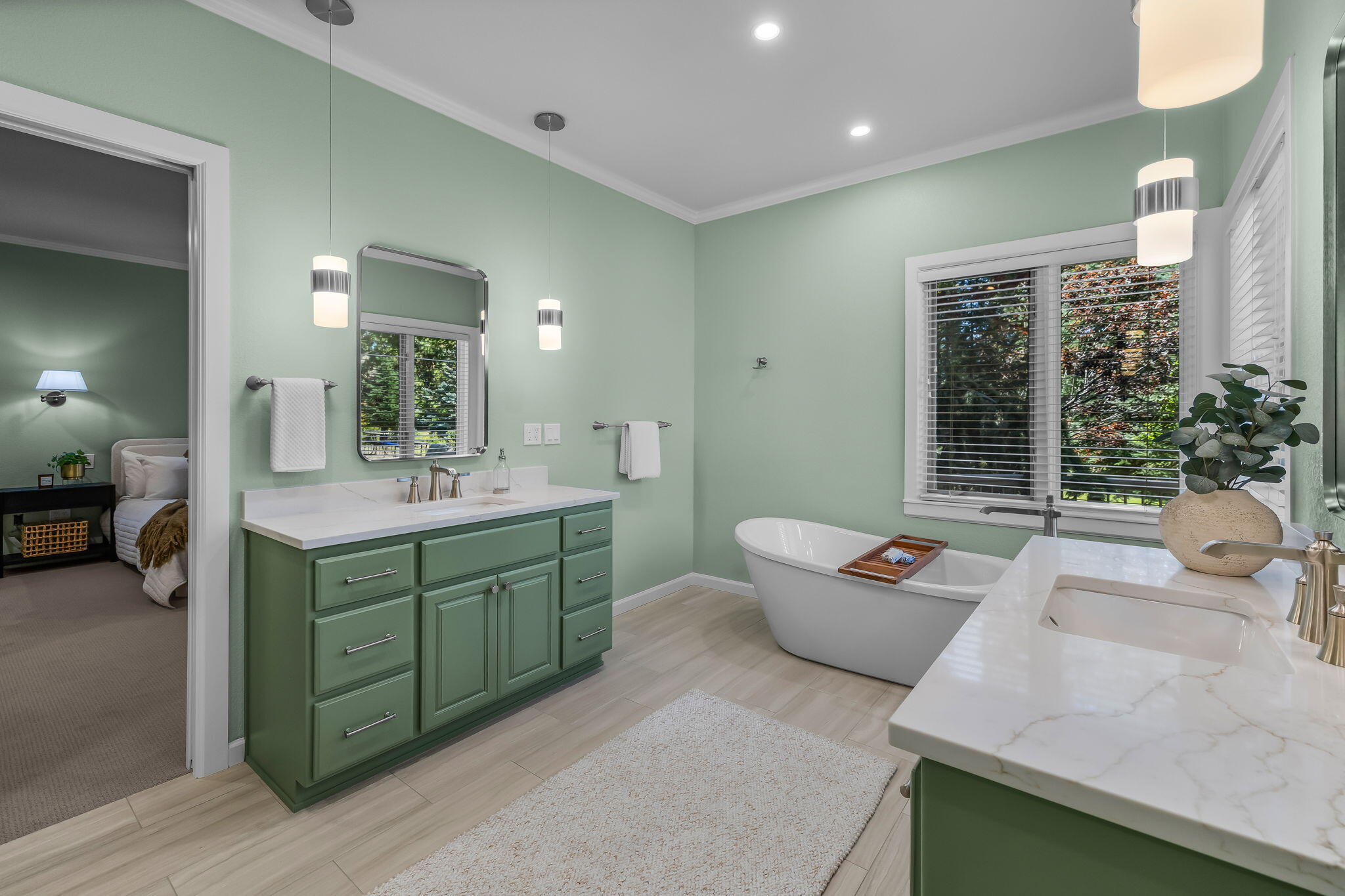 Bathroom with green cabinetry, a freestanding tub, and windows overlooking trees.