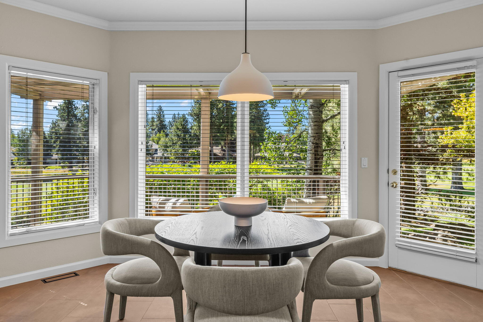 Dining nook with a round table and upholstered chairs beside wide windows overlooking leafy trees.