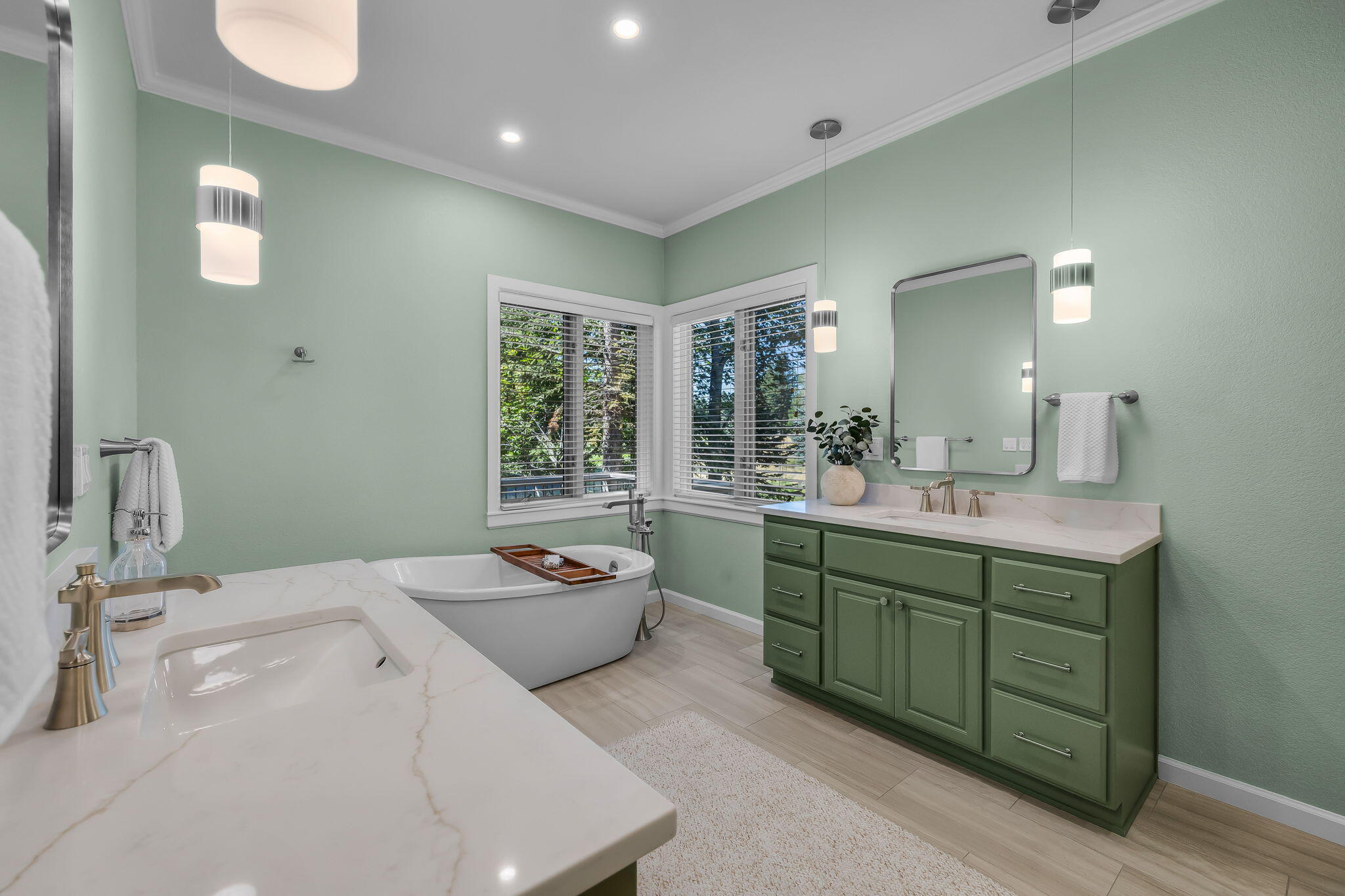 Bathroom with sage-green double vanity, freestanding soaking tub, and corner windows overlooking trees.