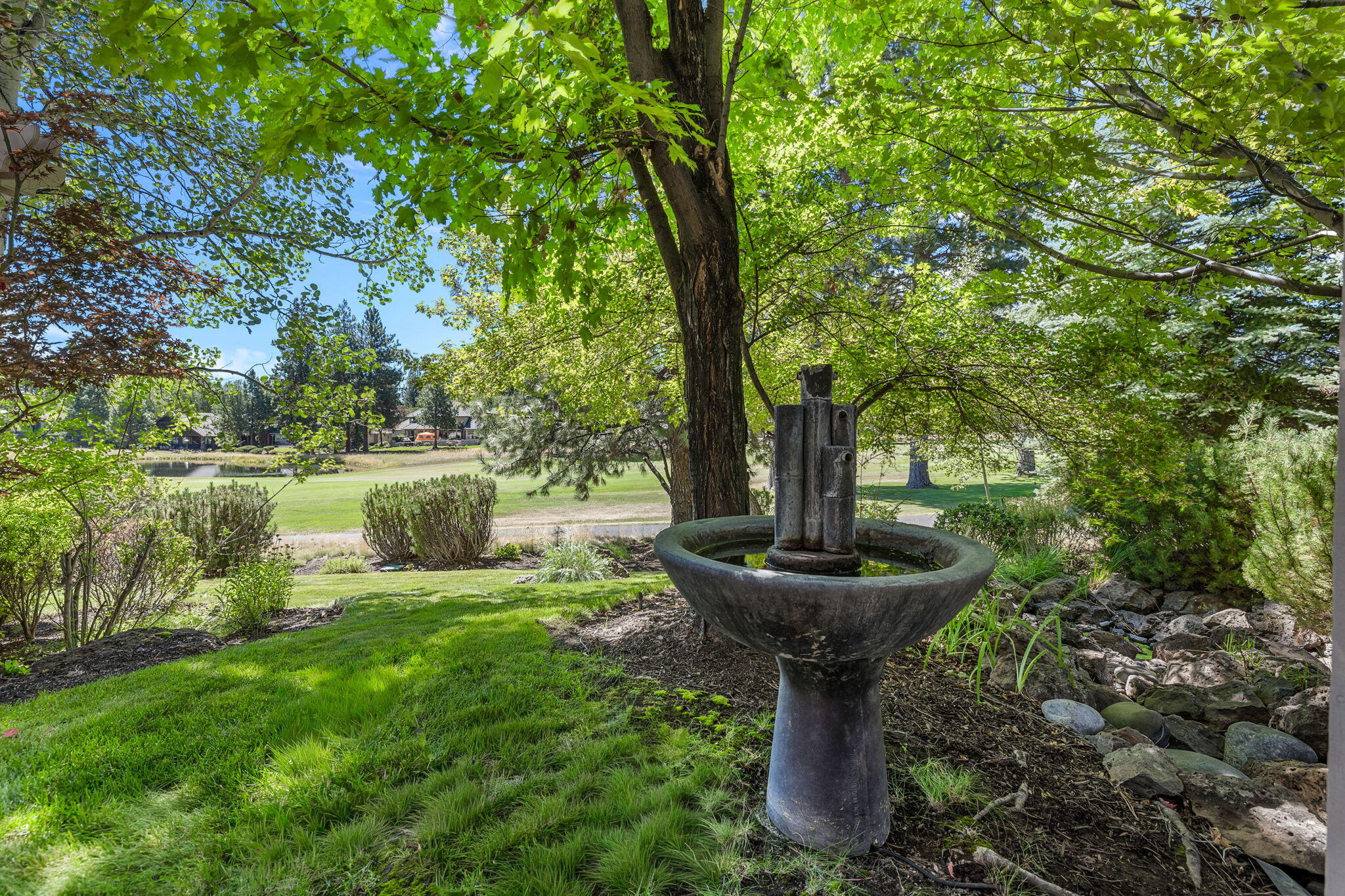 Garden fountain beneath mature trees, with a rock-lined stream and pond beyond the lawn.
