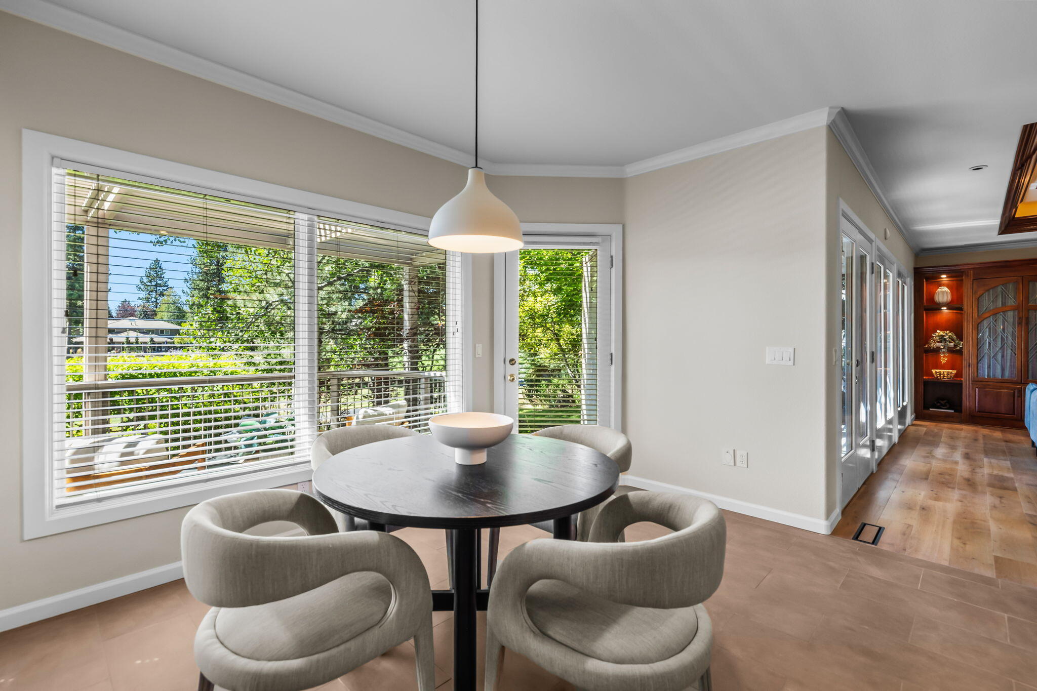 Dining nook with a round table and upholstered chairs beside large windows overlooking trees.