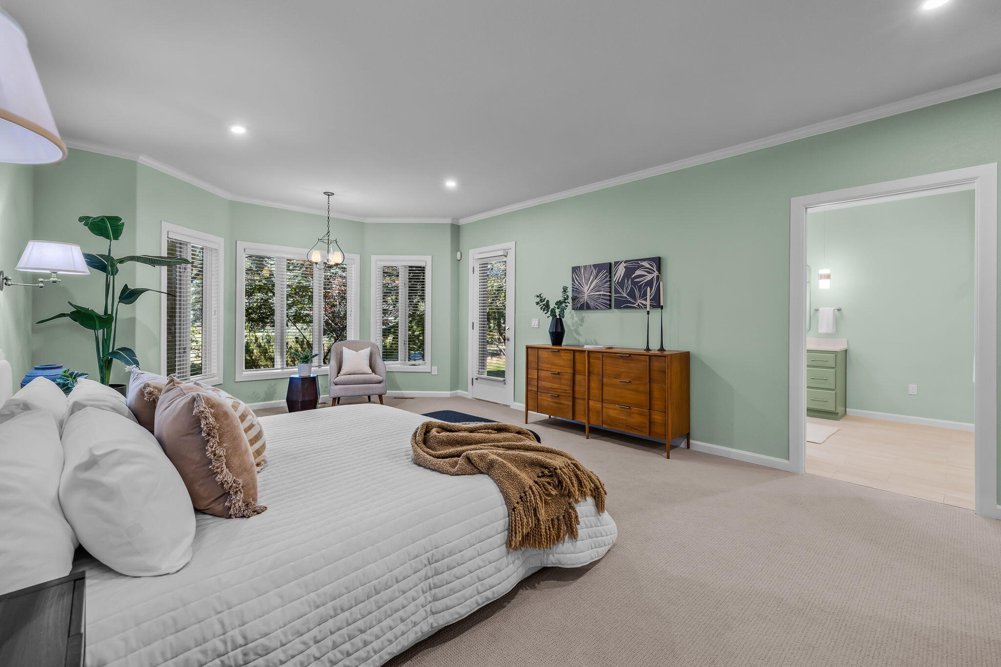 Furnished bedroom with pale green walls, a white bed, wooden dresser, and windows with blinds.
