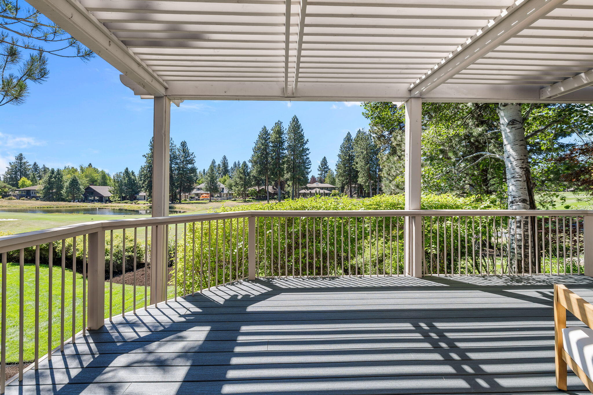 Covered deck overlooking a pond, with white railings, pine trees, and strong striped shadows.