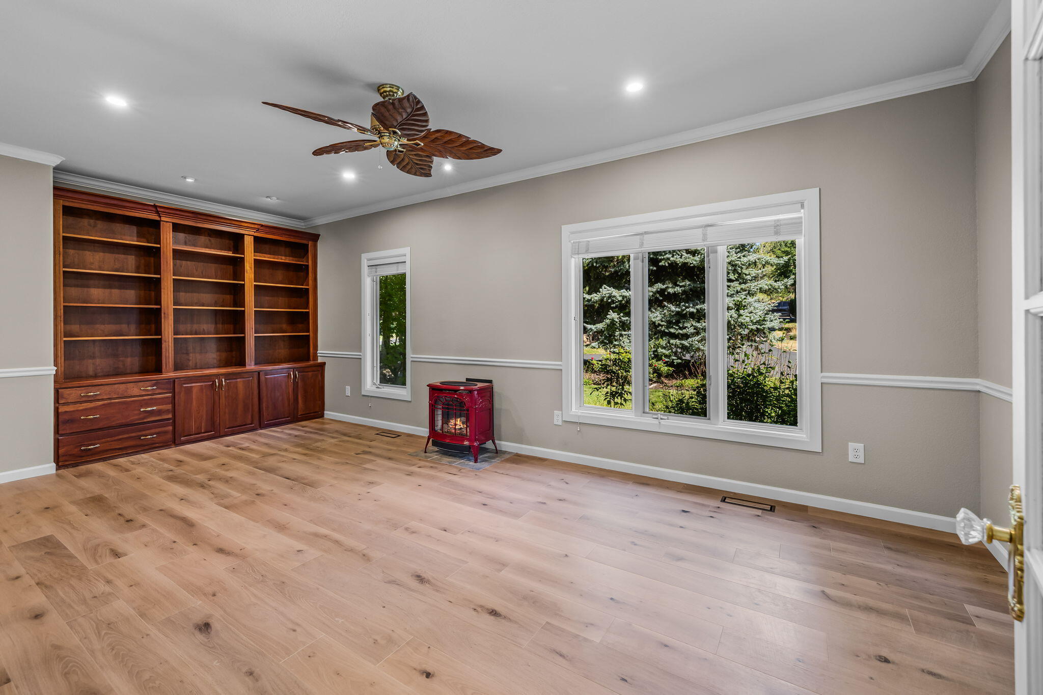 Empty room with built-in wood shelving, a red stove, light wood floors, and windows facing evergreen trees.
