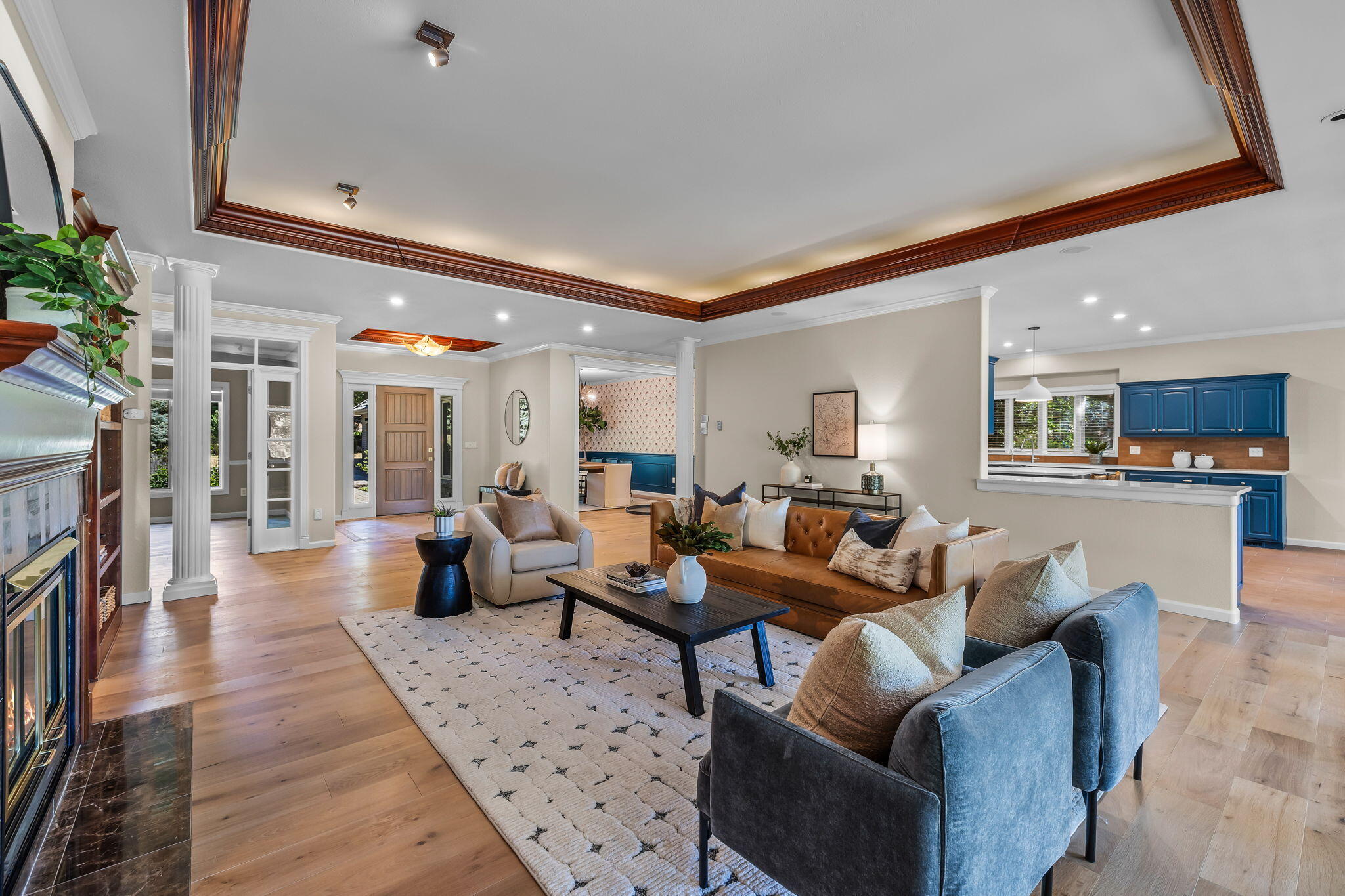 Staged living room with a fireplace, ornate wood-trimmed ceiling, and blue kitchen cabinets.