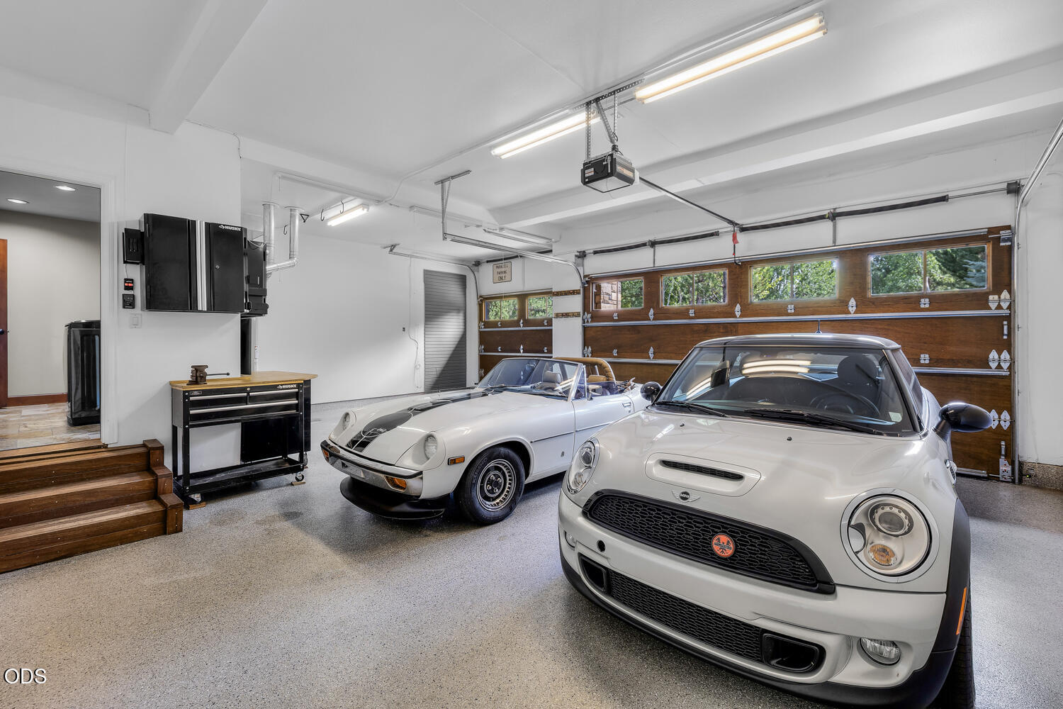 Garage with two white cars, wood-paneled doors, and a rolling tool chest.