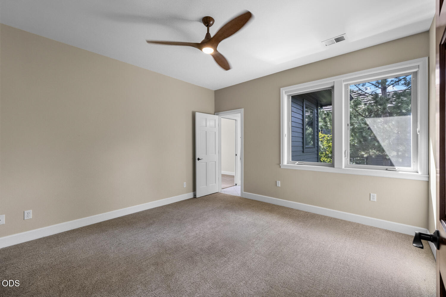 Empty carpeted bedroom with a ceiling fan and large window overlooking trees.