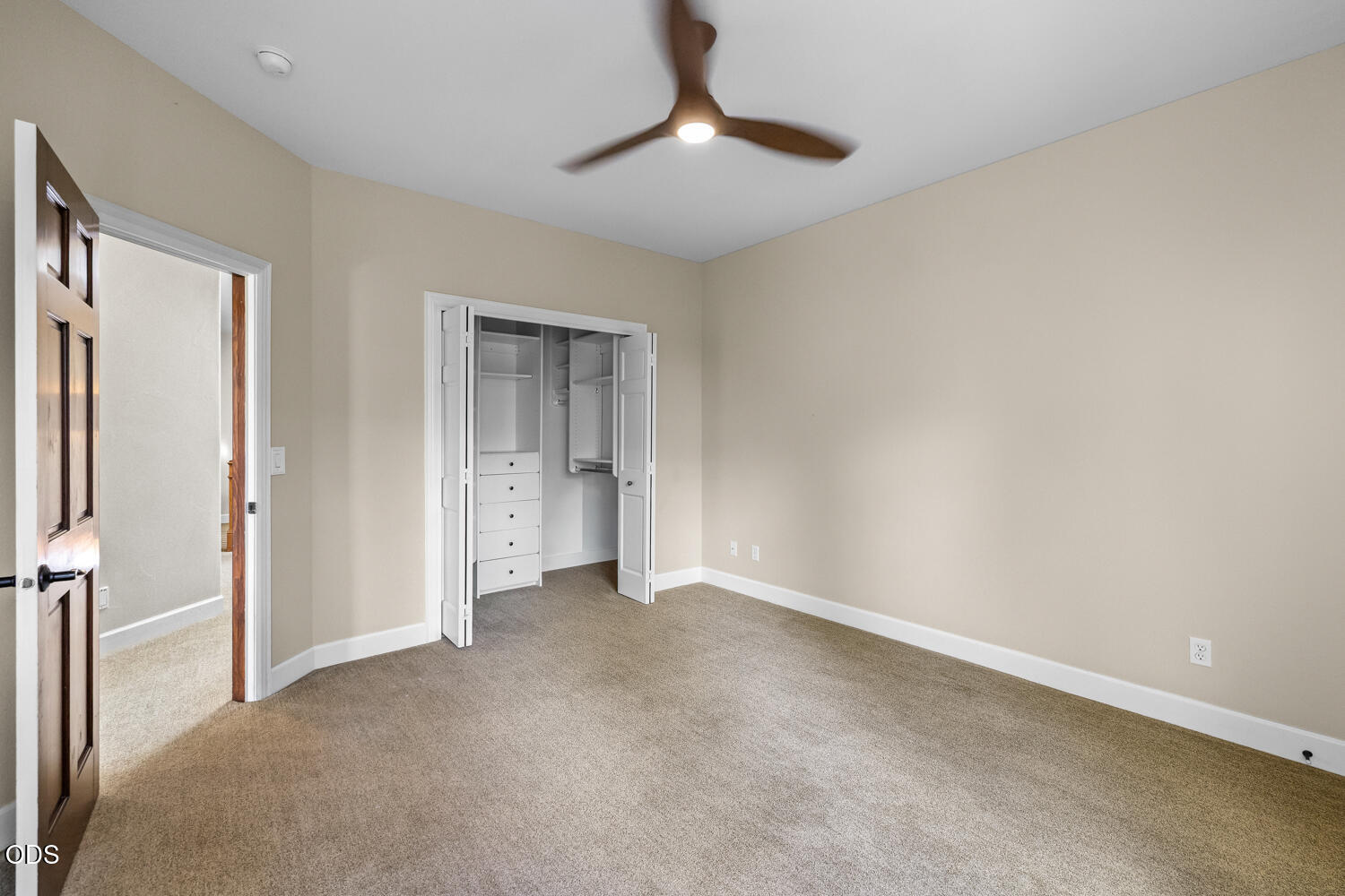 Empty bedroom with tan carpet, ceiling fan, and open closet with built-in drawers.
