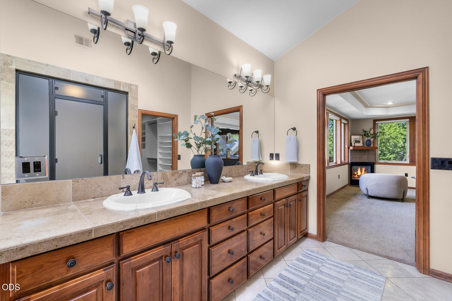 Bathroom with a long double vanity and wood cabinets, opening to a sitting room with a fireplace.
