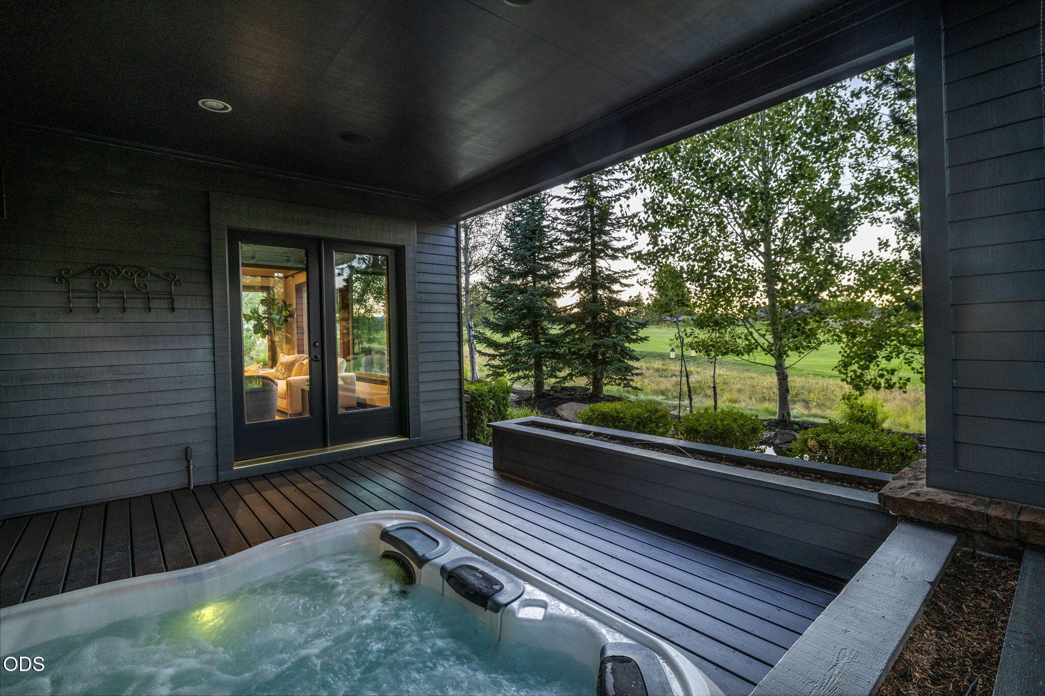 Covered patio with a built-in hot tub overlooking a landscaped lawn and mature trees.