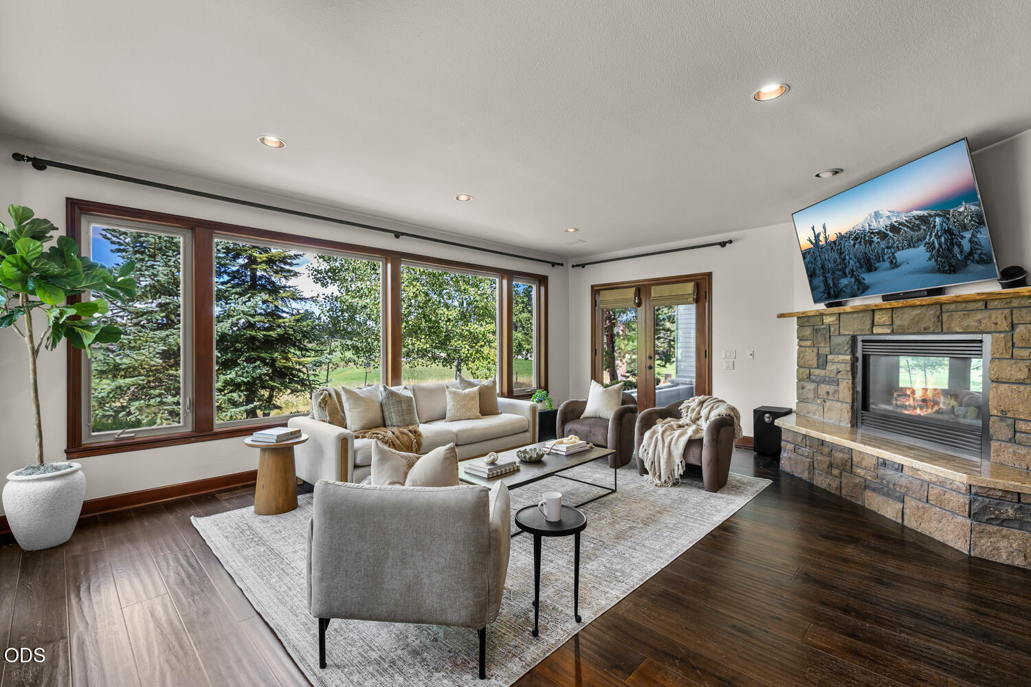 Living room with large windows overlooking trees and a stone fireplace with a television.