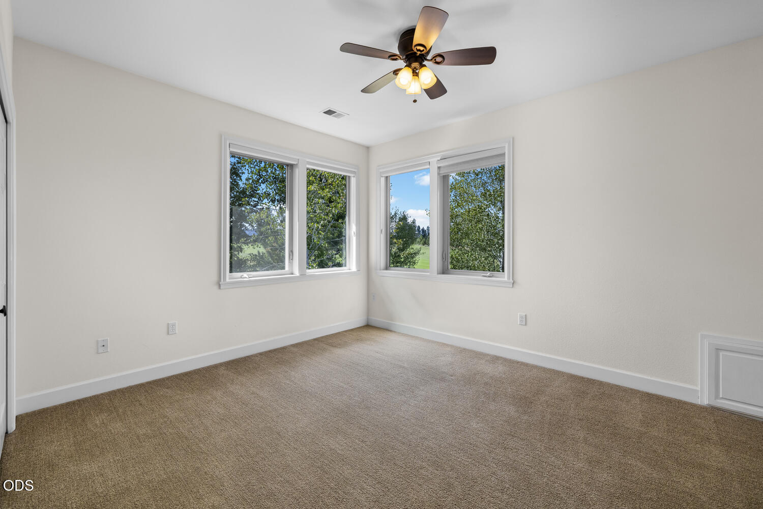 Empty carpeted room with a ceiling fan and corner windows overlooking trees and lawn.