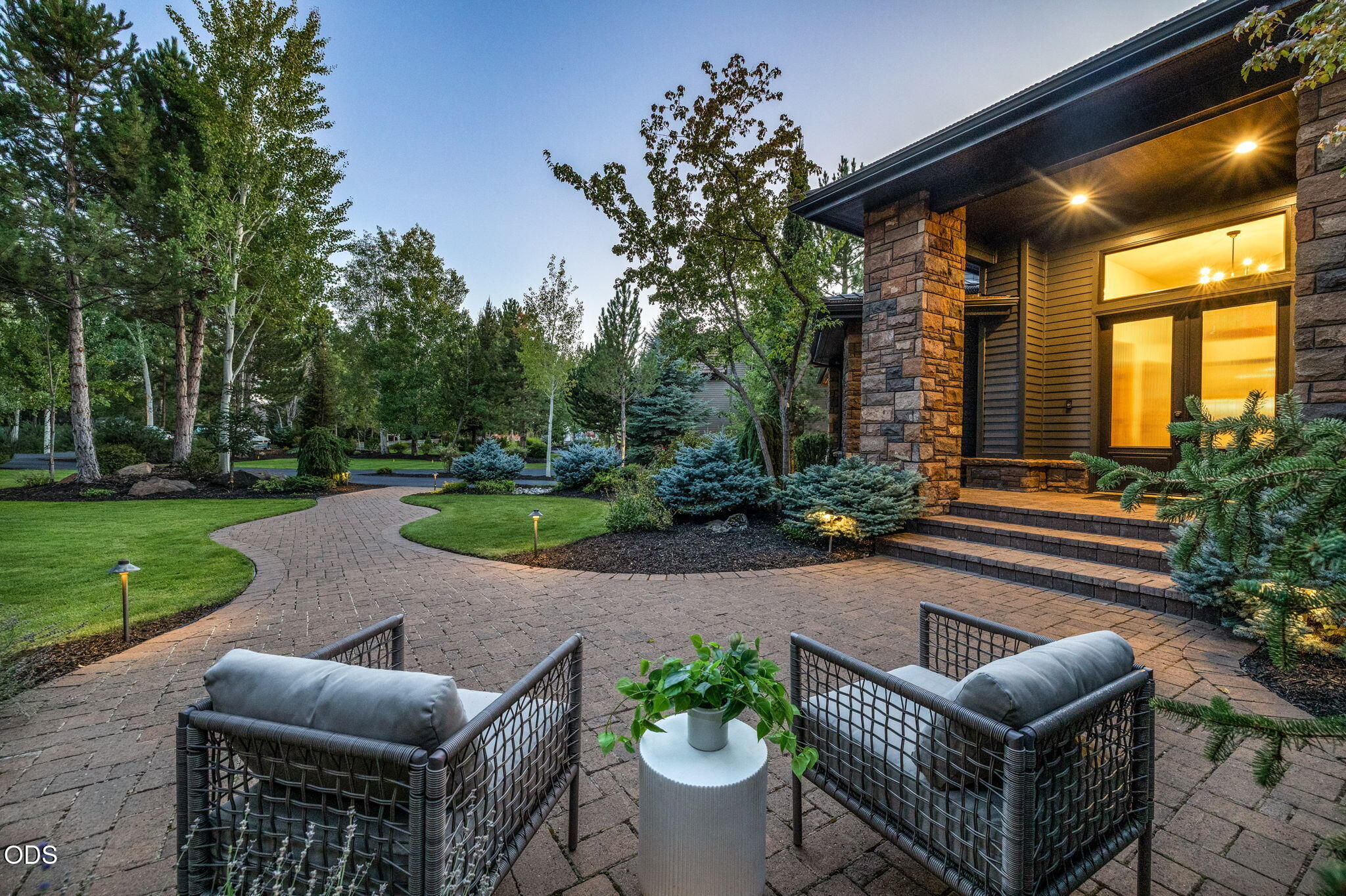 Furnished brick patio beside a stone-clad home, with cushioned chairs and a curved garden walkway.