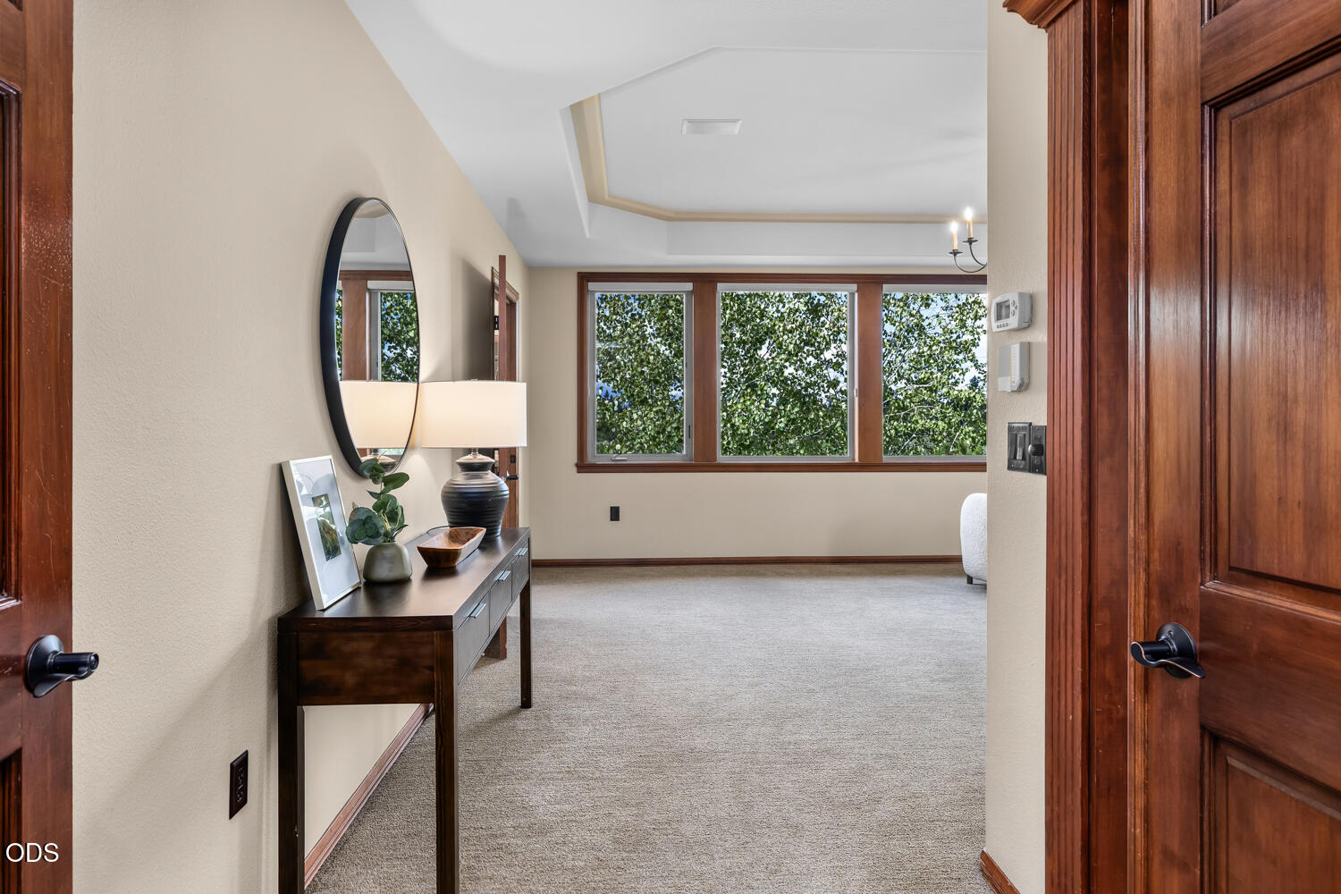 Carpeted room with large wood-trimmed windows, leafy views, and a dark wood console table by the doorway.