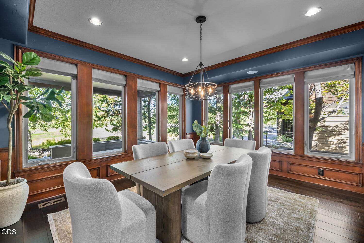 Dining room with wood-trimmed windows, blue walls, and a long table surrounded by upholstered chairs.