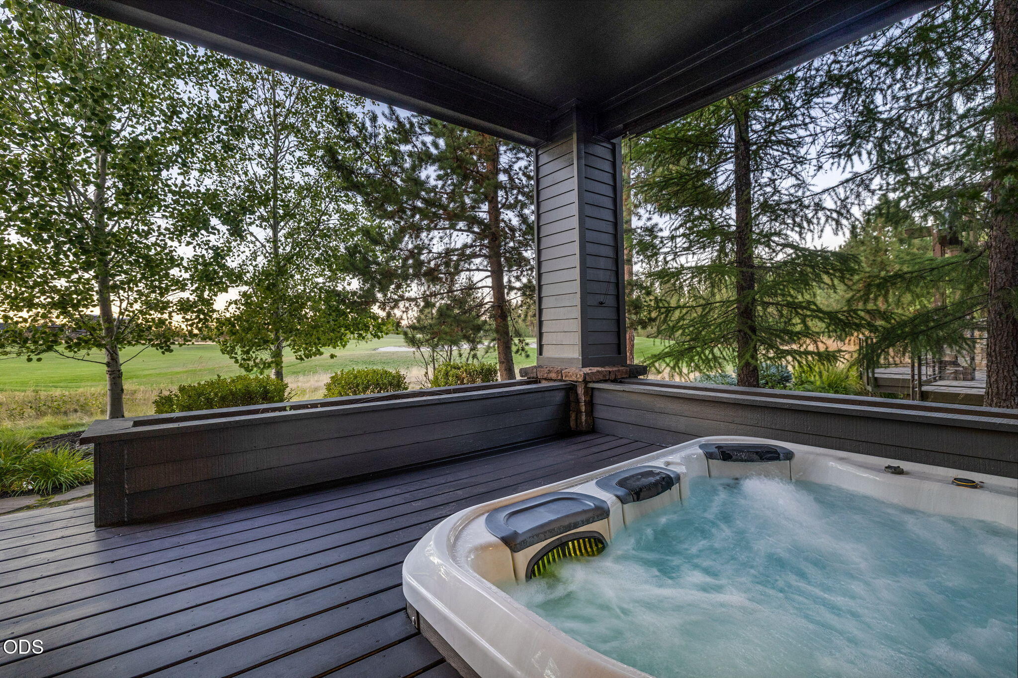 Covered deck with a bubbling hot tub overlooking a manicured green and trees.