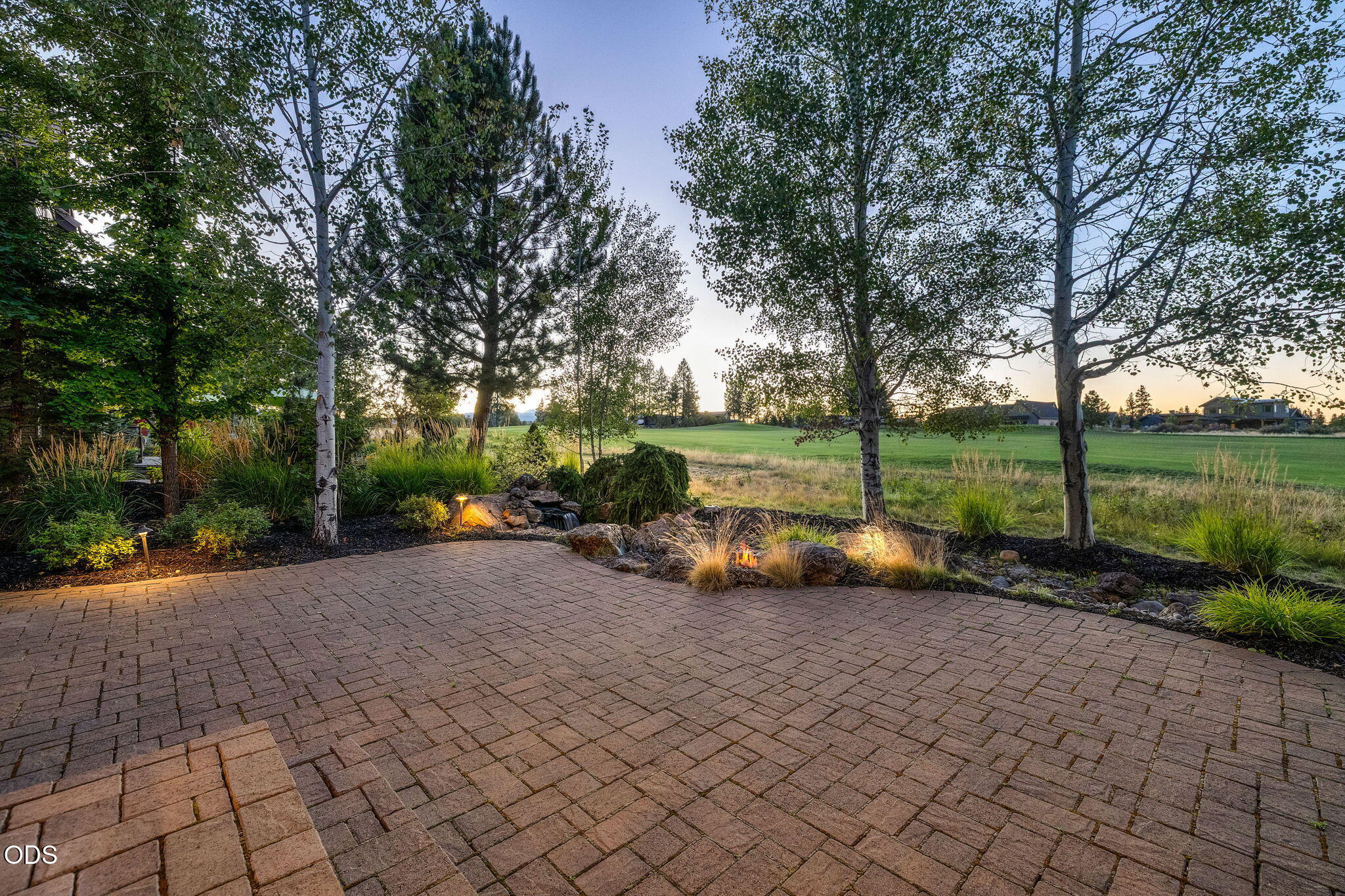Brick-paver patio with landscaped beds and a small water feature overlooking an open green lawn.