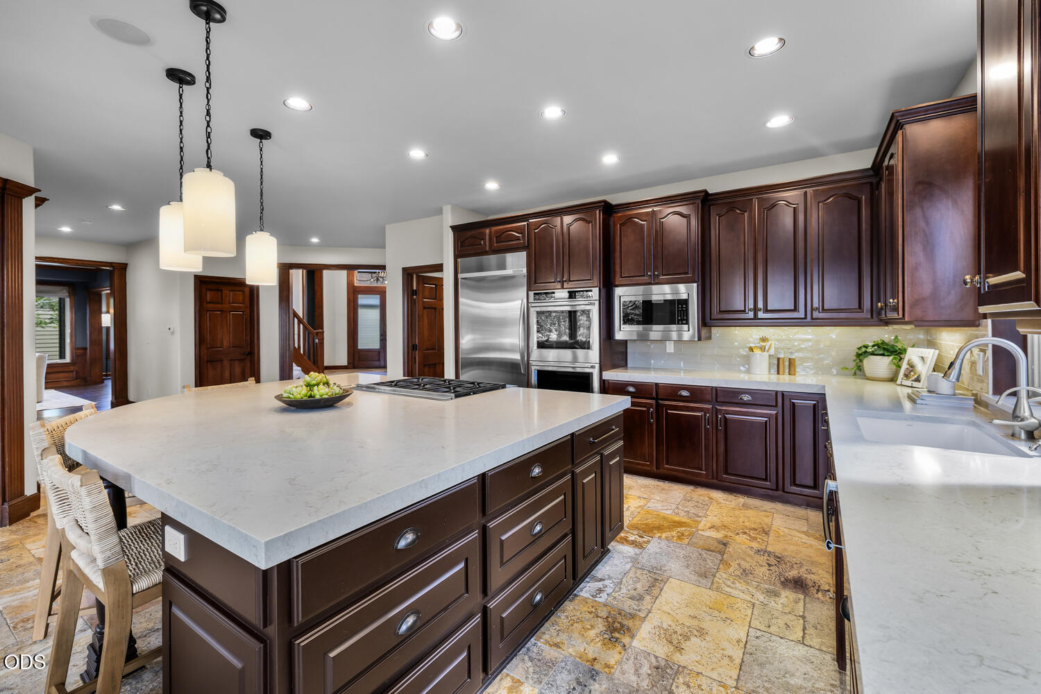 Kitchen with a large white island, dark cherry cabinets, stainless appliances, and stone tile flooring.