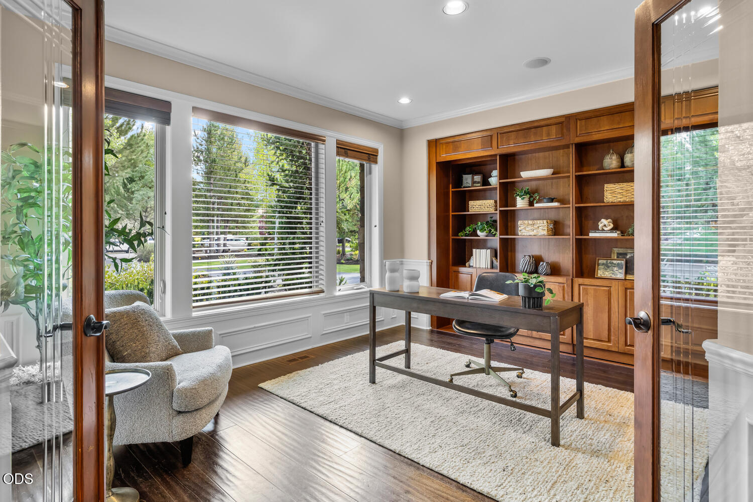 Furnished home office with built-in wooden bookcases, a desk, and wide windows overlooking greenery.