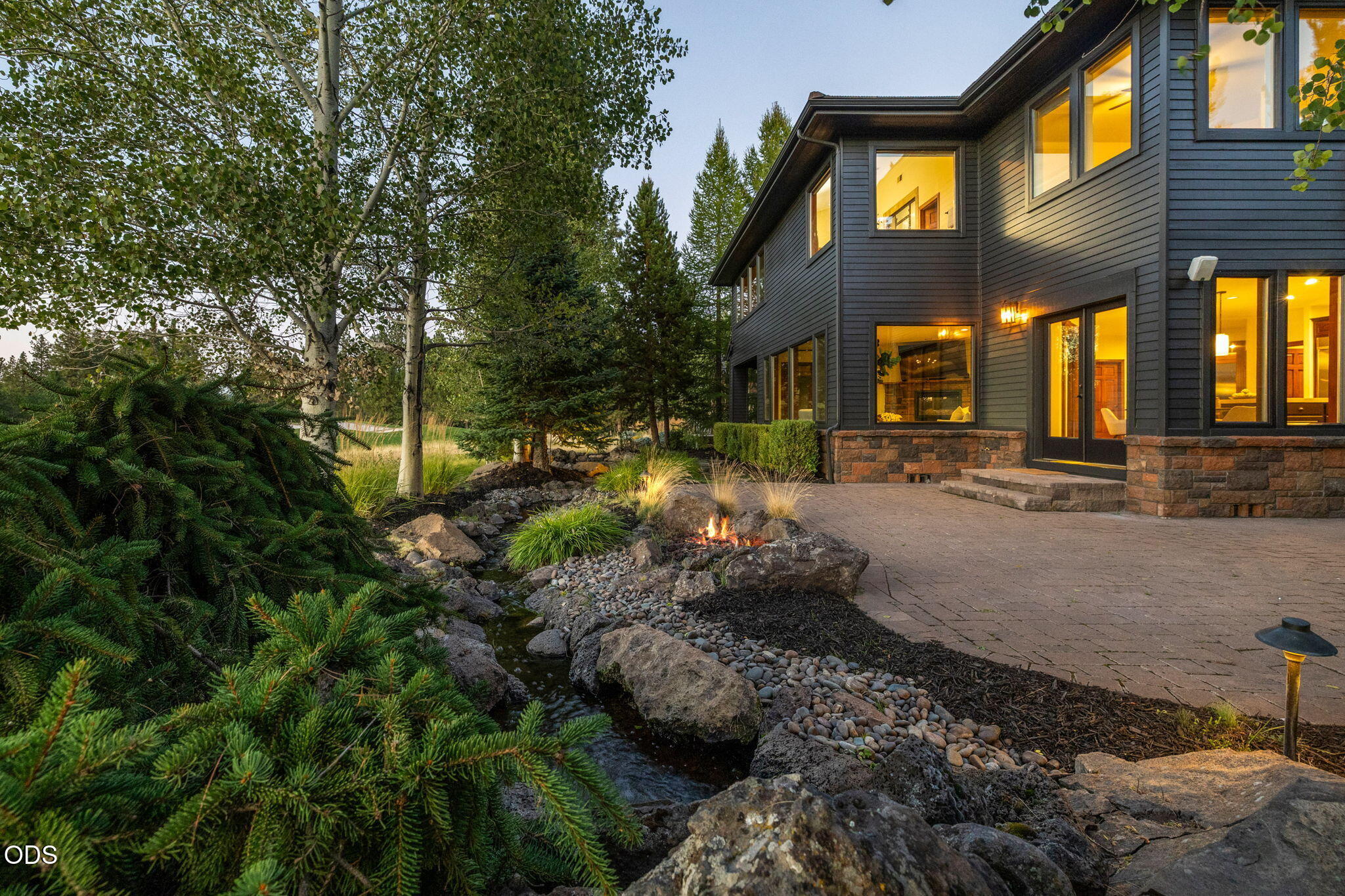 Modern two-story home beside a landscaped rock stream and small fire feature, with warmly lit windows at dusk.