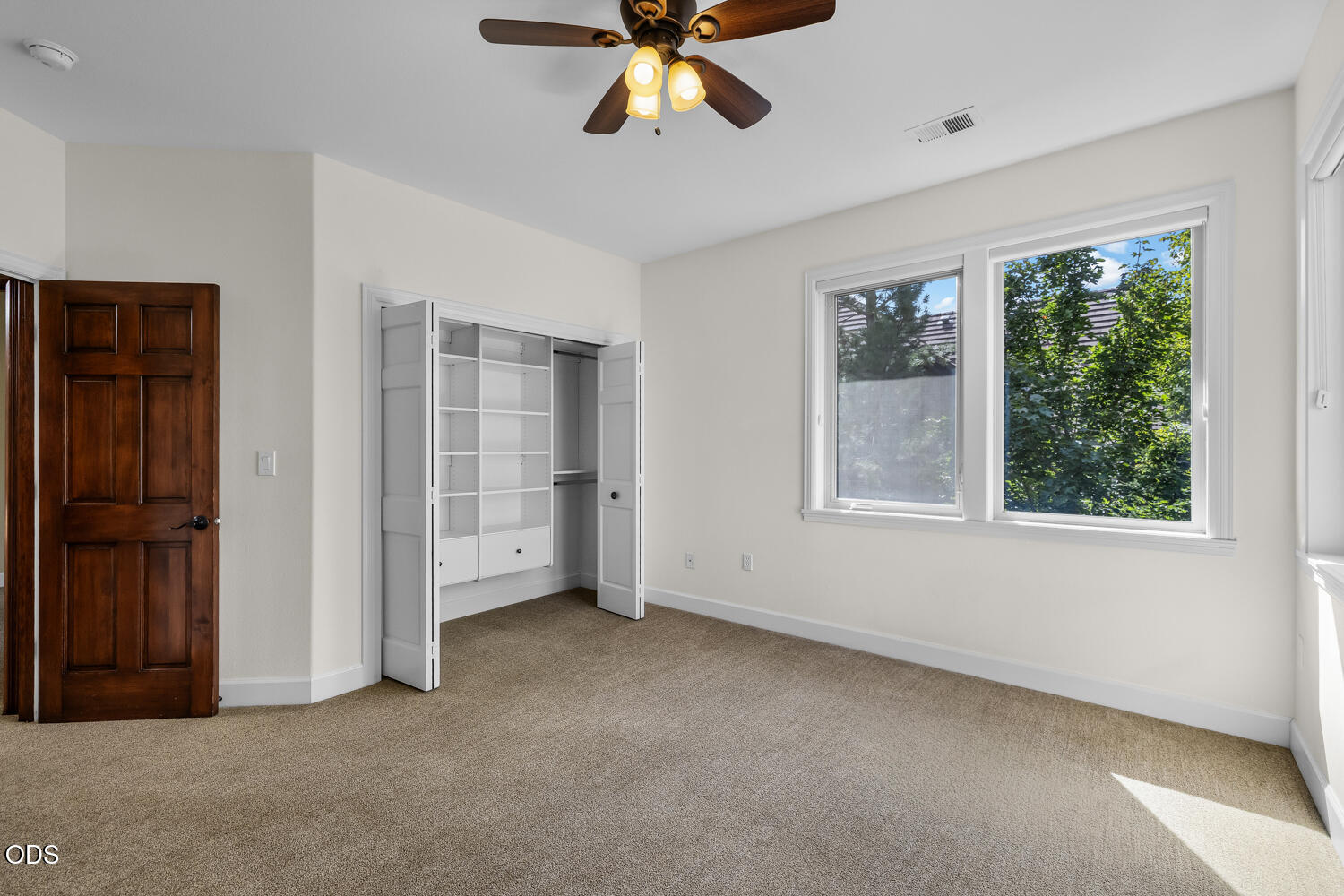 Empty bedroom with an open built-in closet, ceiling fan, and large windows overlooking greenery.