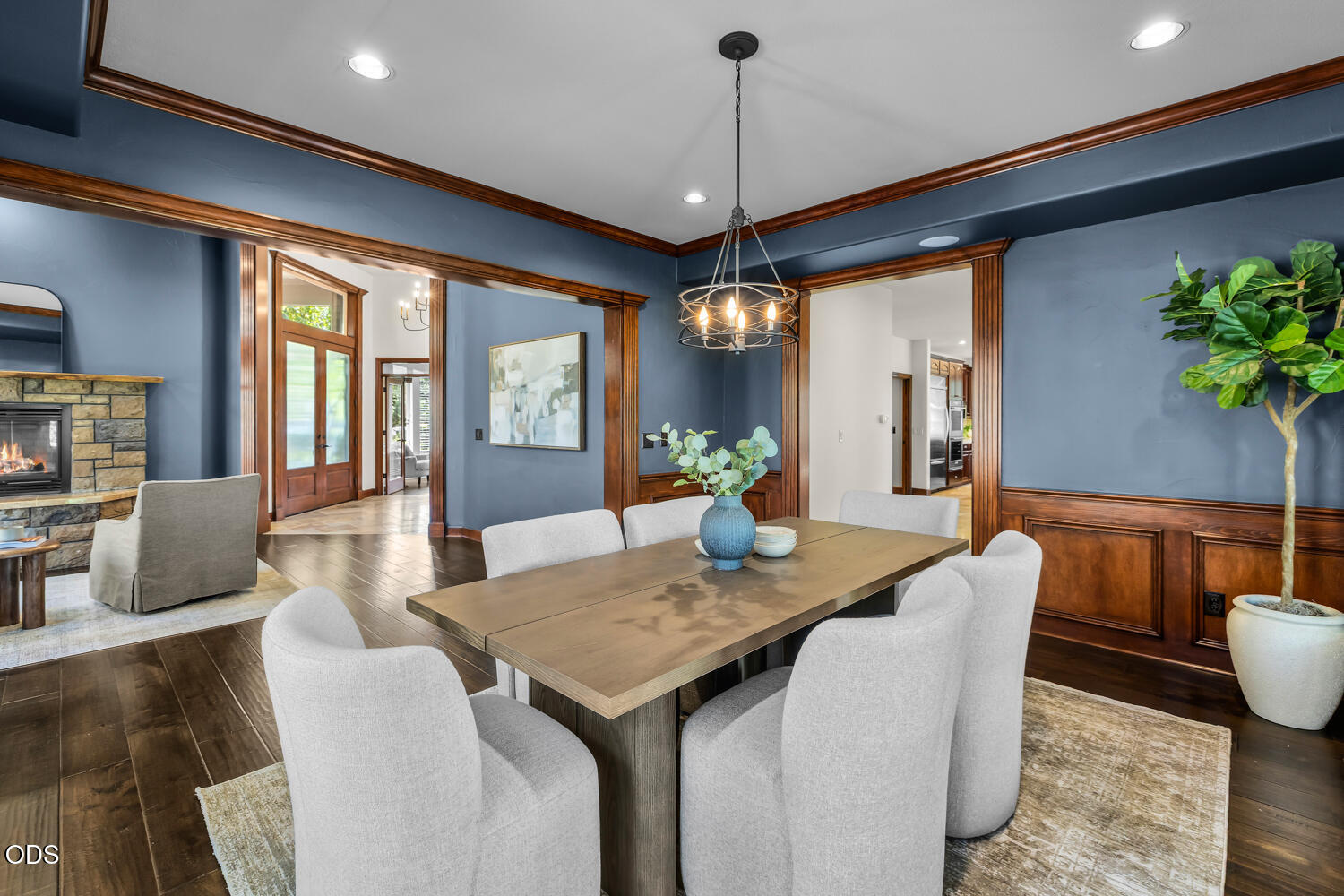Dining room with a long table, blue walls, wood trim, and a chandelier beside a stone fireplace.