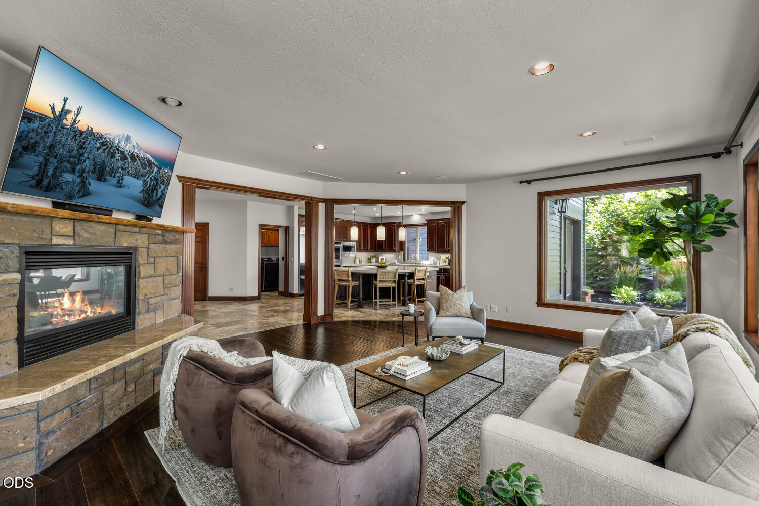 Furnished living room with a stone fireplace, large window, and open kitchen beyond.