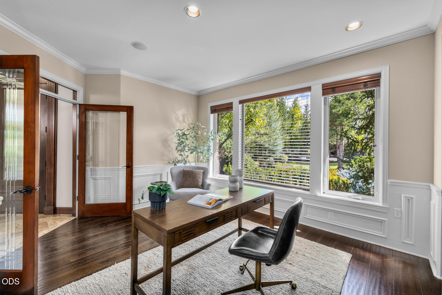 Home office with a wooden desk, black chair, and large windows overlooking leafy greenery.
