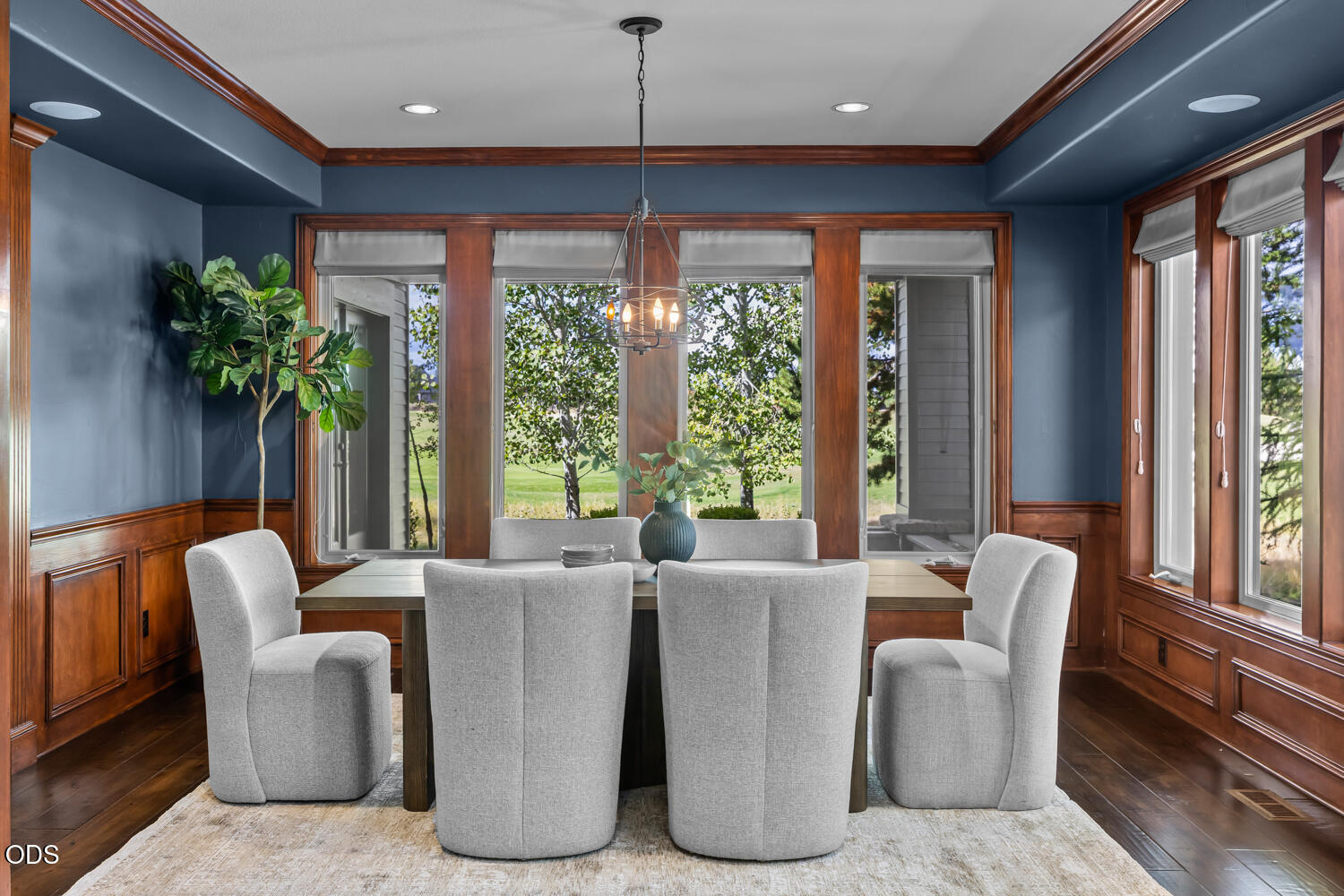 Dining room with gray upholstered chairs, wood trim, and large windows overlooking a green lawn.