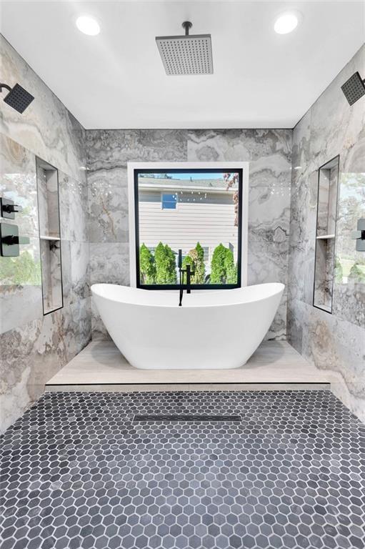 Bathroom with a freestanding tub beneath a window, marble walls, and hexagonal tile flooring.