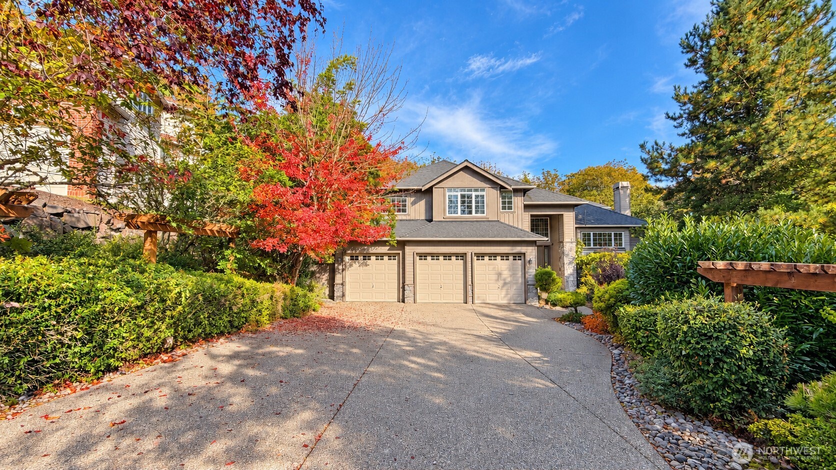 Two-story beige home with a three-car garage, broad driveway, and red autumn foliage.