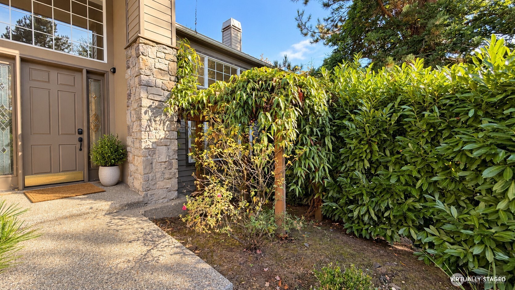 Home entrance with a paneled door, stone facade, paved walkway, and dense hedges.