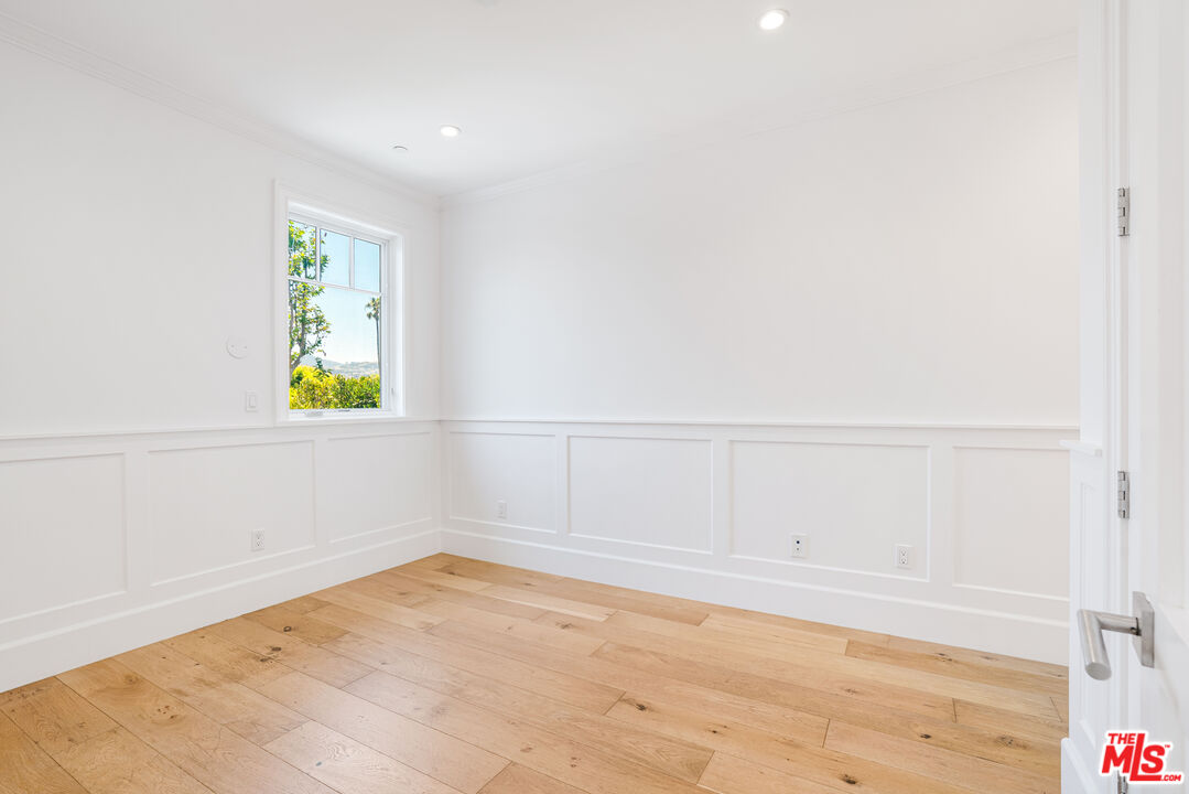 Empty room with white panel molding, wide-plank wood floors, and a window overlooking greenery.