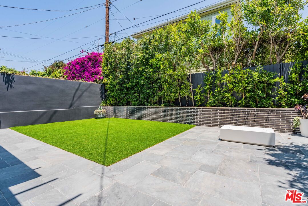 Backyard patio with gray stone paving, a green lawn, white bench, and dense privacy landscaping.