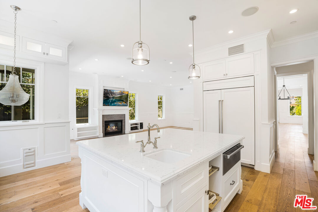 Bright white kitchen with a marble island and farmhouse sink, opening to a living area with a fireplace.