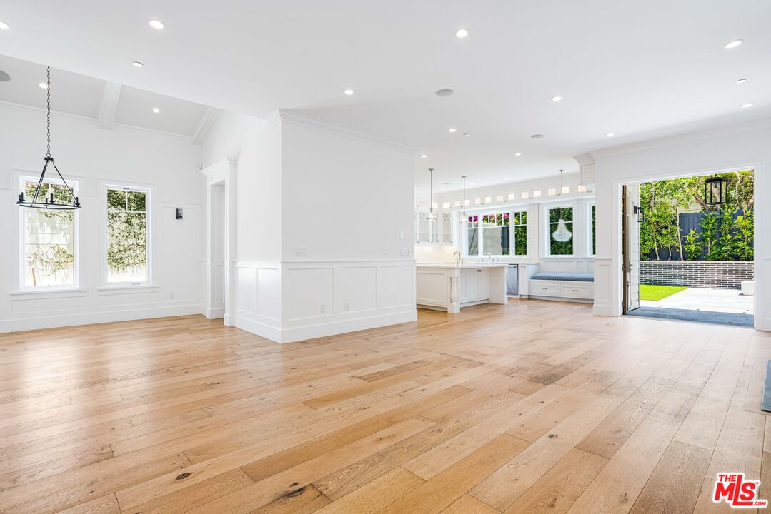 Vacant open-plan room with wide-plank wood floors, white paneling, and a kitchen opening onto a patio.