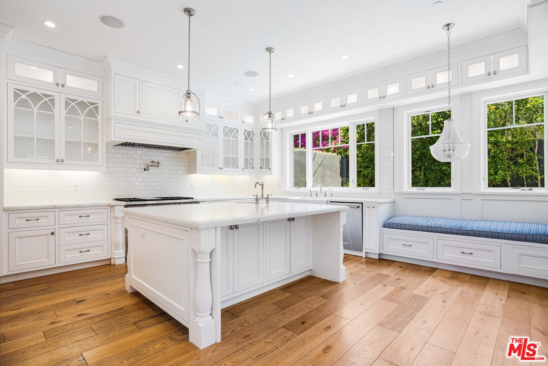 Bright white kitchen with a large island, glass-front cabinets, warm wood floors, and windows overlooking greenery.