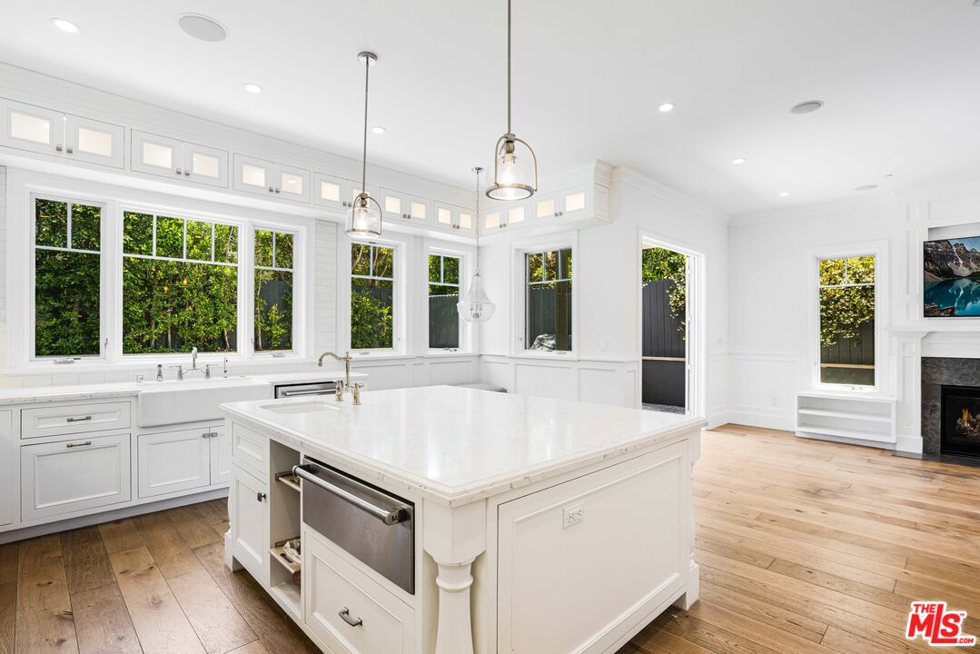 White kitchen with a large marble island, long windows overlooking greenery, and hardwood floors.