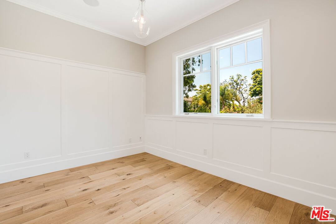 Bright empty room with oak floors, white paneled walls, and a large window overlooking trees.
