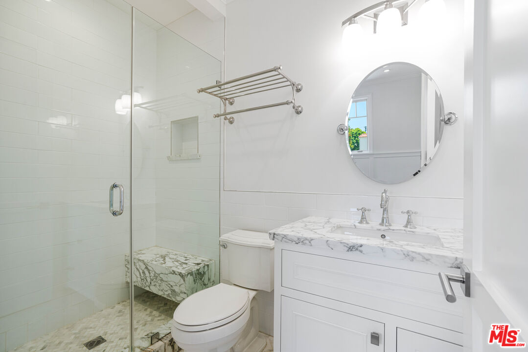 White bathroom with a glass shower, marble-patterned bench, and marble vanity beneath a round mirror.