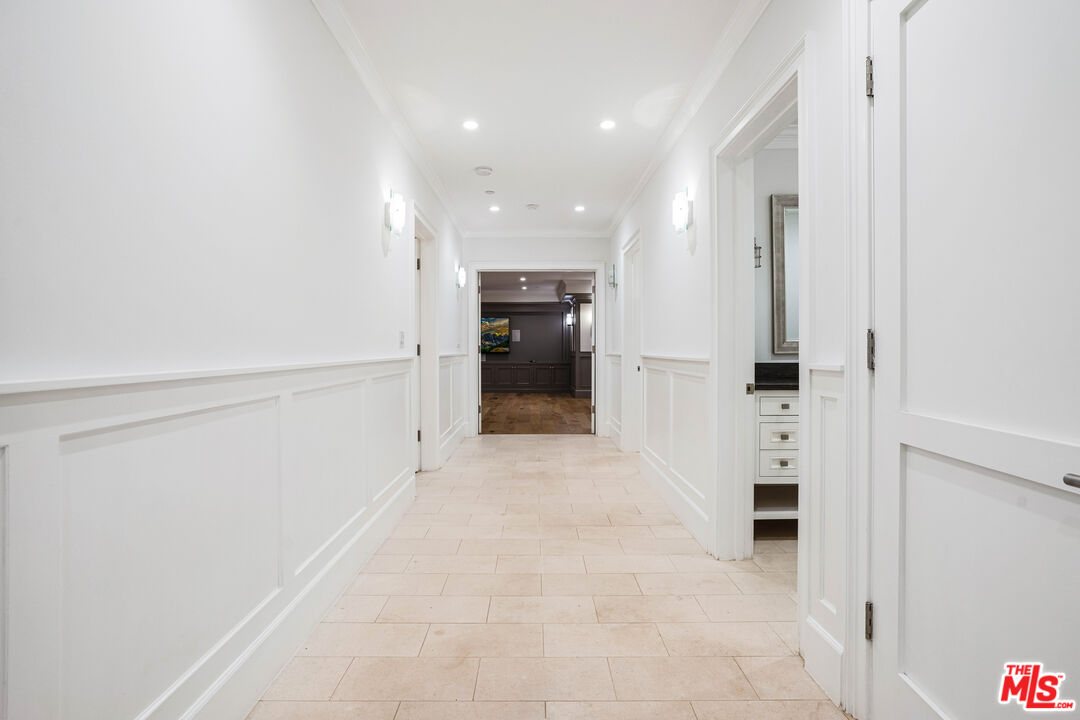 White paneled hallway with pale tile floors, recessed lights, and a built-in vanity visible through a doorway.