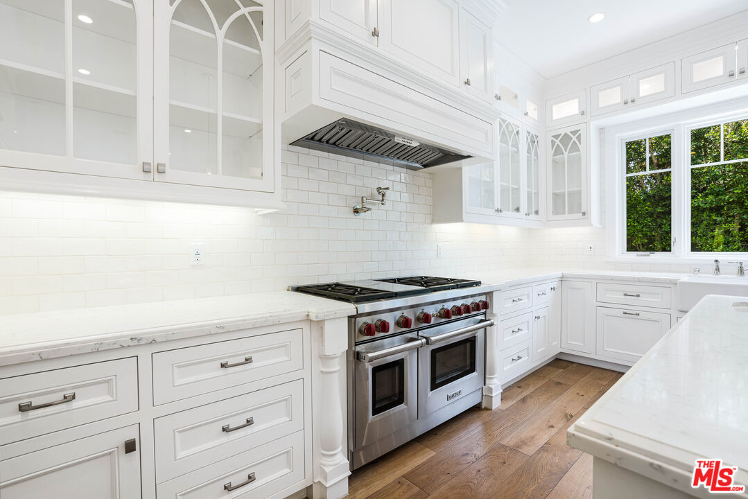White kitchen with glass-front cabinetry, marble counters, a stainless steel range, and windows overlooking greenery.