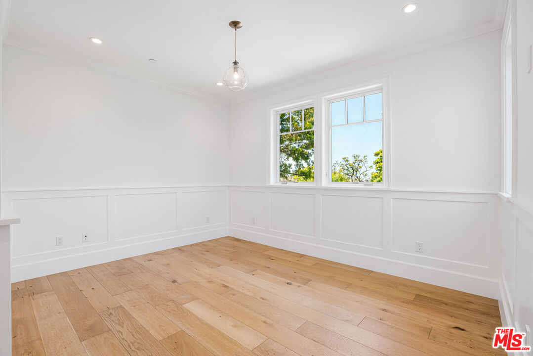 Vacant room with white paneled walls, wide-plank wood floors, and windows overlooking leafy trees.