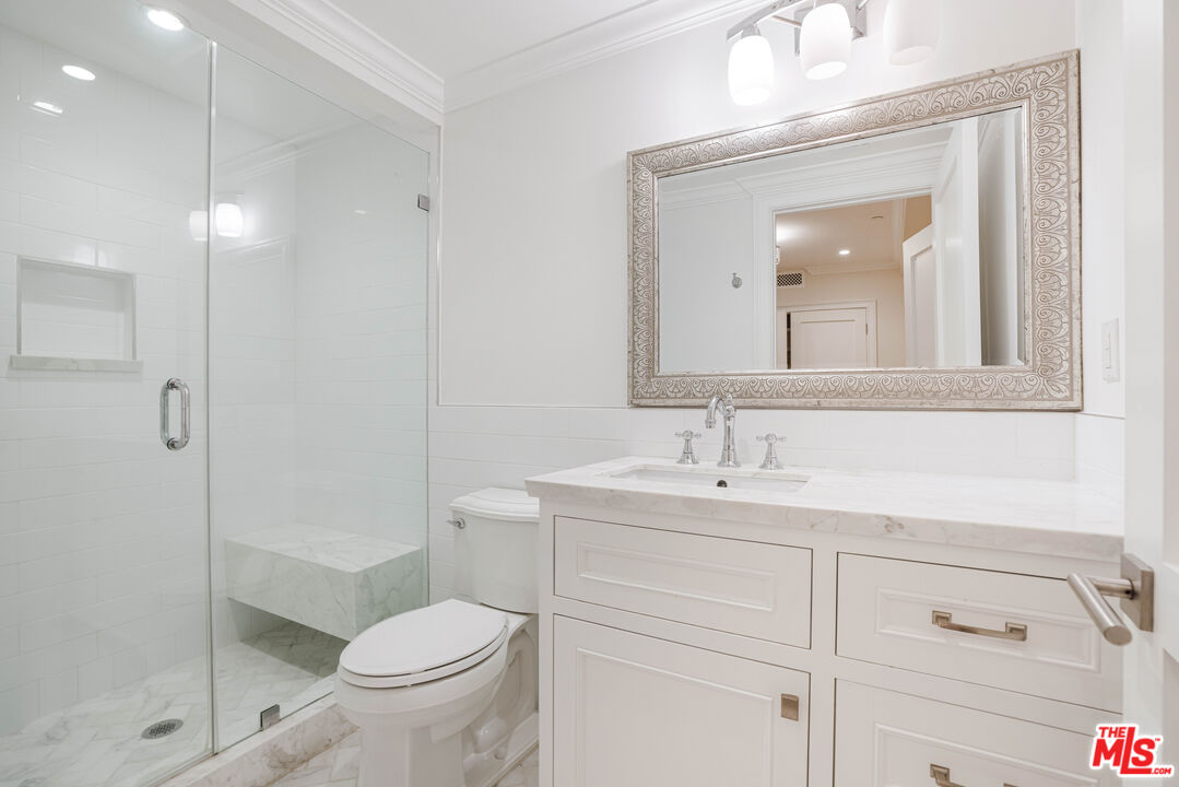 White bathroom with a glass shower, marble bench, and vanity beneath an ornate mirror.