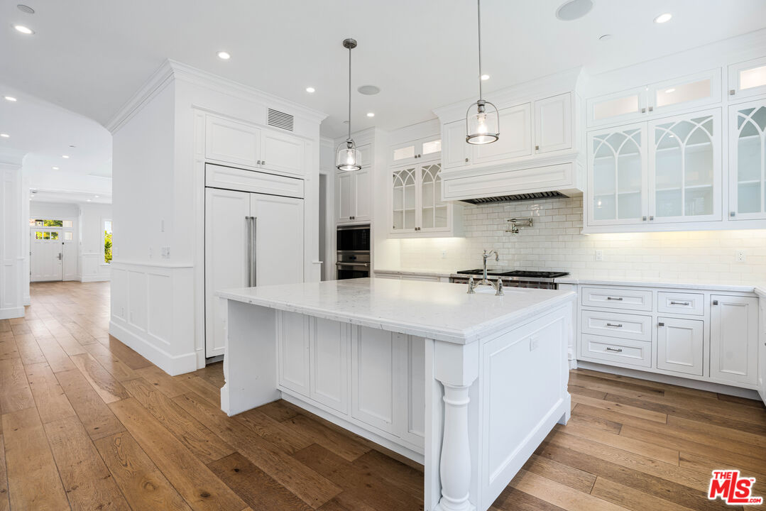 White kitchen with a large marble island, glass-front cabinets, and wide-plank wood floors.