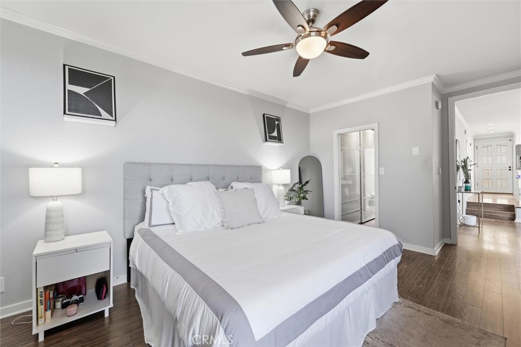 Staged bedroom with a white bed, gray upholstered headboard, ceiling fan, and dark wood flooring.