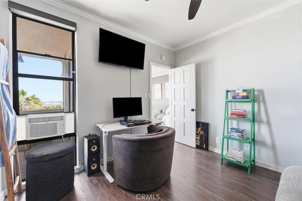Furnished media room with computer screens, wall-mounted TV, green bookshelf, and window air conditioner.