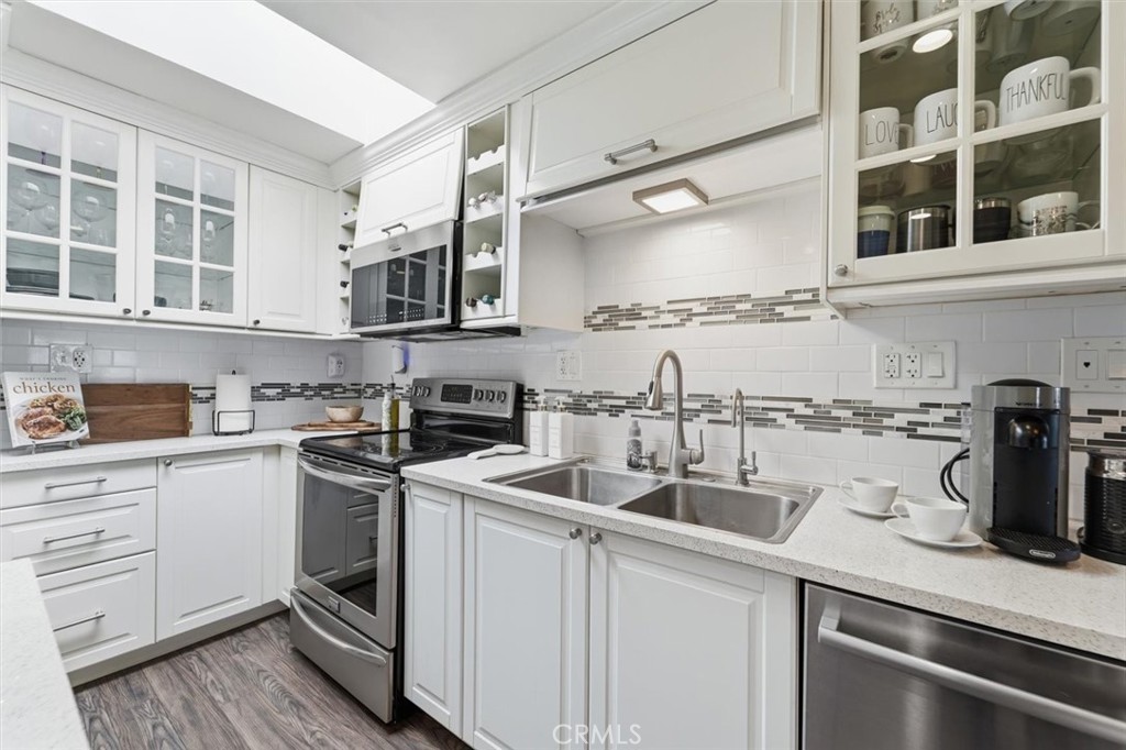 White kitchen with stainless appliances, a double-basin sink, glass-front cabinets, and mosaic backsplash.