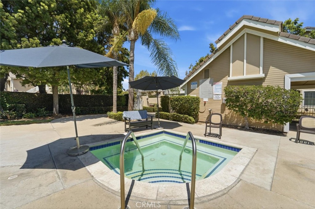 A tiled square spa with metal handrails sits on a sunny patio beside umbrellas and a tan building.
